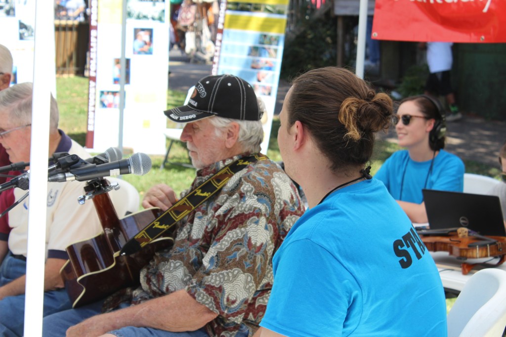Three men in the foreground play string instruments while Ellie, in the background, monitors sound equipment
