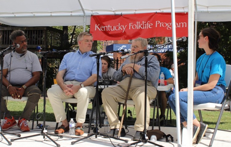 Three older men and Ellie sit on a stage with microphones. A banner behind them reads "Kentucky Folklife Program"